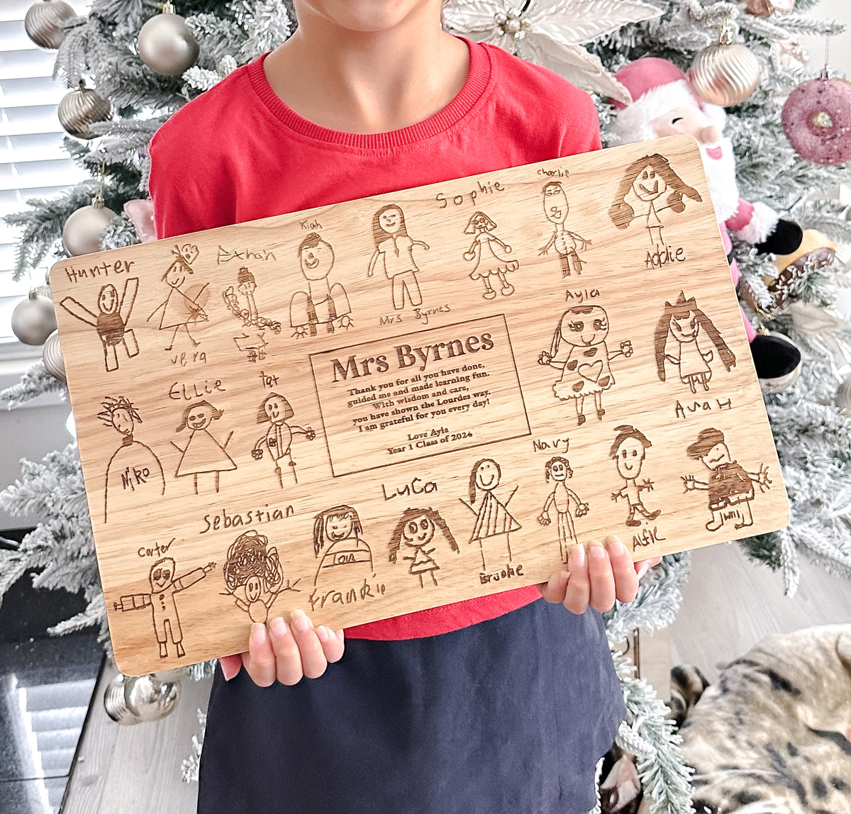 Person holding a wooden board with hand-drawn family illustrations and text for a teacher end of year gift, in front of a decorated Christmas tree.