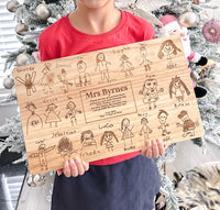 Person holding a wooden board with hand-drawn family illustrations and text for a teacher end of year gift, in front of a decorated Christmas tree.